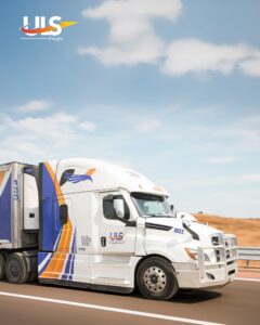 A white semi-truck with blue and orange stripes and a bird logo, branded “UIS Freight,” driving on a highway through a desert landscape under a blue sky with scattered clouds.