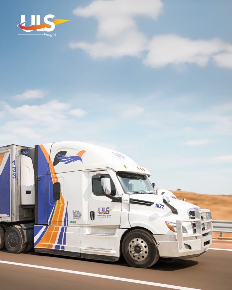A white semi-truck with blue and orange stripes and a bird logo, branded “UIS Freight,” driving on a highway through a desert landscape under a blue sky with scattered clouds.