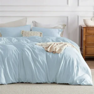 Soft light blue bedding set styled on a neatly made bed, featuring matching pillowcases, a textured cream accent pillow, and a folded beige throw blanket. The bed sits against a white paneled wall with a wooden nightstand to the side, creating a calm, airy bedroom aesthetic.