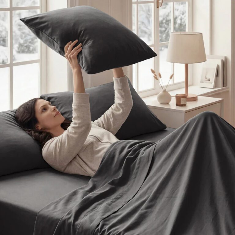 Woman lying in a neatly made bed with dark gray sheets, holding a matching pillow above her face. Soft natural light comes through large windows behind her, and a bedside table with a lamp and small decor adds to the calm, cozy bedroom setting.