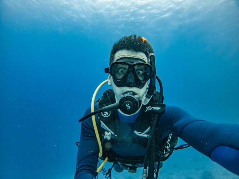 A diver taking a selfie underwater with a surprised expression, wearing a wetsuit and diving mask.